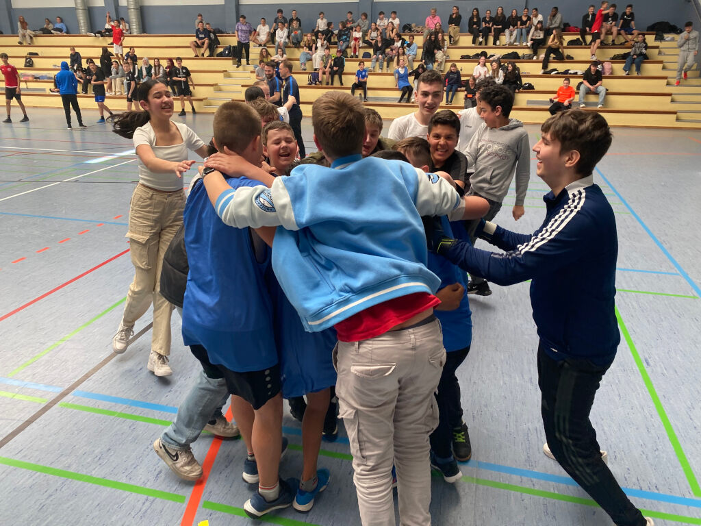 Eine Gruppe Jugendlicher jubelt und umarmt sich ausgelassen auf einem Hallenboden; im Hintergrund sitzen Zuschauende auf den Rängen.