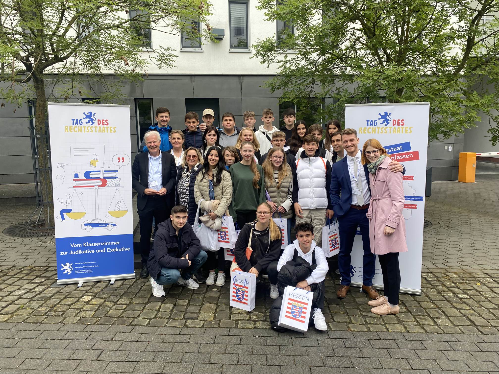 Eine Schulklasse posiert mit mehreren Erwachsenen im Freien zwischen zwei Roll-ups mit der Aufschrift „Tag des Rechtsstaates“; einige halten Taschen mit dem Hessen-Logo.