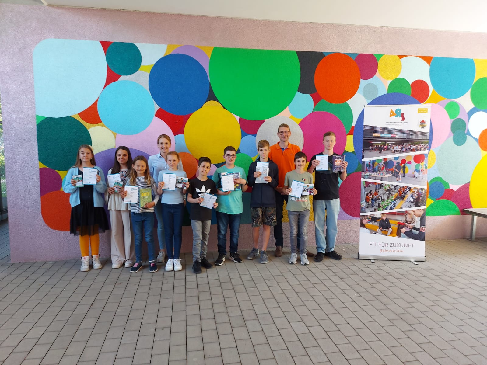 Eine Gruppe Kinder hält Urkunden und kleine Preise in die Kamera, dazu zwei Lehrkräfte, vor einer bunt mit Kreisen bemalten Wand neben einem ARS-Roll-up.