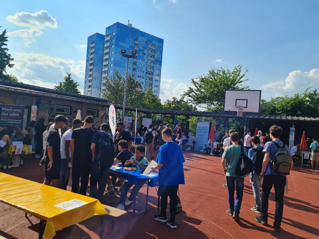 Eine Berufsinfo-Veranstaltung im Freien auf einem Sportplatz mit Basketballkorb: Jugendliche gehen zwischen Informationsständen mit Bannern wie „Duales Studium – Ausbildung“ umher; im Hintergrund ein blaues Hochhaus.