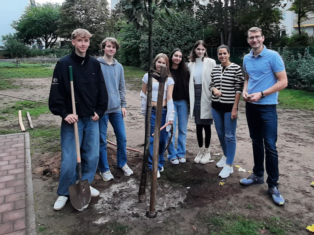 Eine Gruppe Jugendlicher steht mit zwei Erwachsenen um einen frisch gepflanzten jungen Baum mit Stützpfahl; eine Person hält einen Spaten.