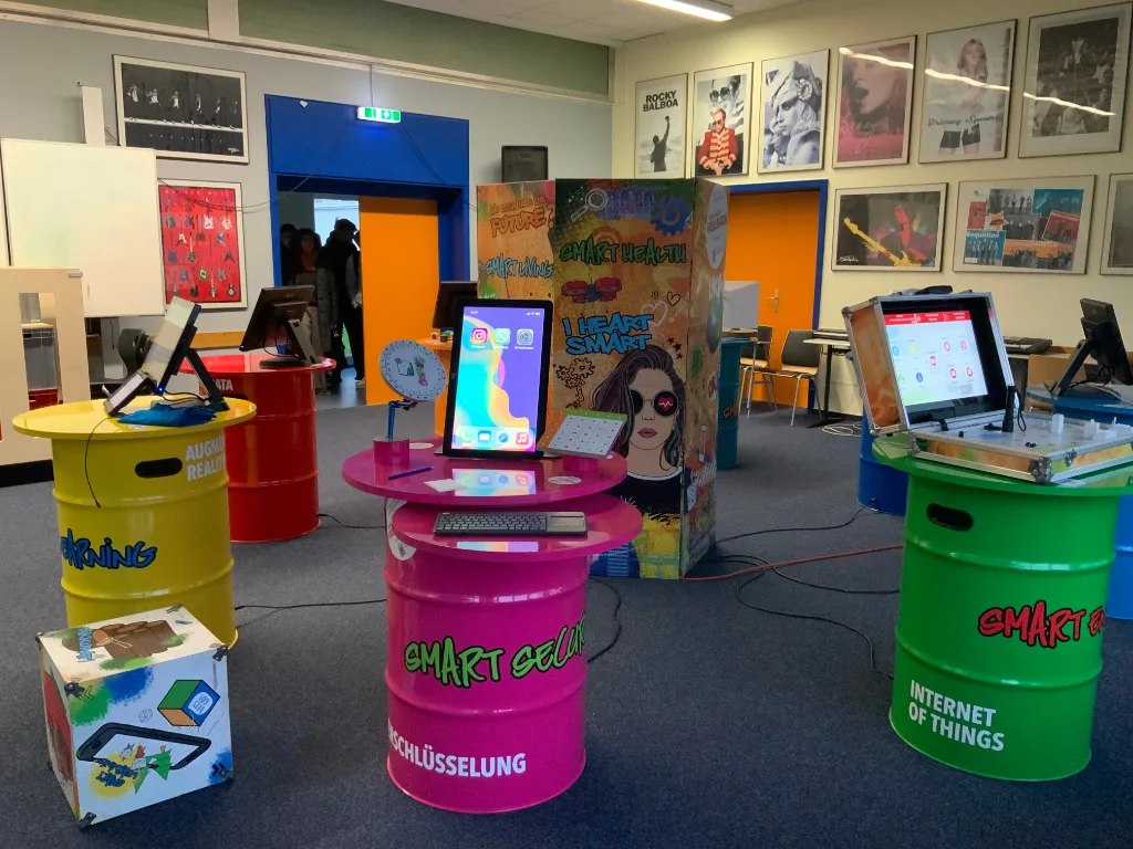 Bunt lackierte Fässer dienen als Lernstationen mit Tablets und Bildschirmen, beschriftet unter anderem mit „Verschlüsselung“ und „Internet of Things“.