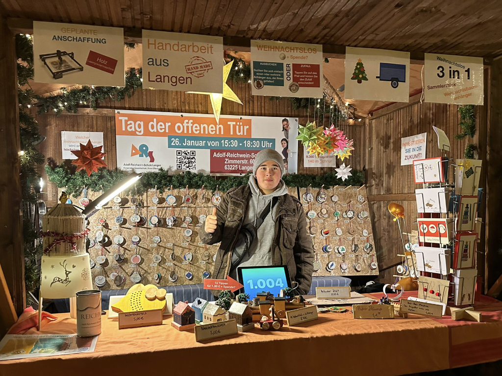 Eine jugendliche Person zeigt mit erhobenem Daumen hinter einem weihnachtlich beleuchteten Verkaufsstand voller selbst gebastelter Holzdeko, Karten und Anhänger; an der Rückwand Plakate der Adolf-Reichwein-Schule.