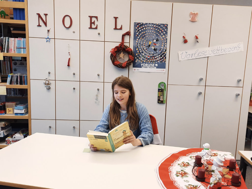 Ein Kind liest an einem Tisch in der Schulbücherei aus einem Buch vor; an der Wand dahinter weihnachtliche Deko mit den Buchstaben „NOEL“ und einem „Vorlesewettbewerb“-Schriftzug.