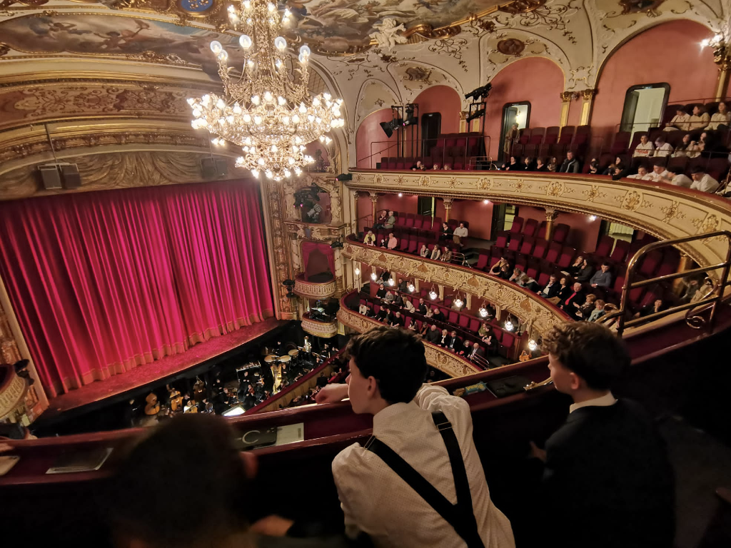 Blick von einem oberen Rang über den prunkvollen Zuschauerraum des Staatstheaters mit rotem Bühnenvorhang, goldverzierten Rängen, Kronleuchter und Orchestergraben; im Vordergrund einige Jugendliche.