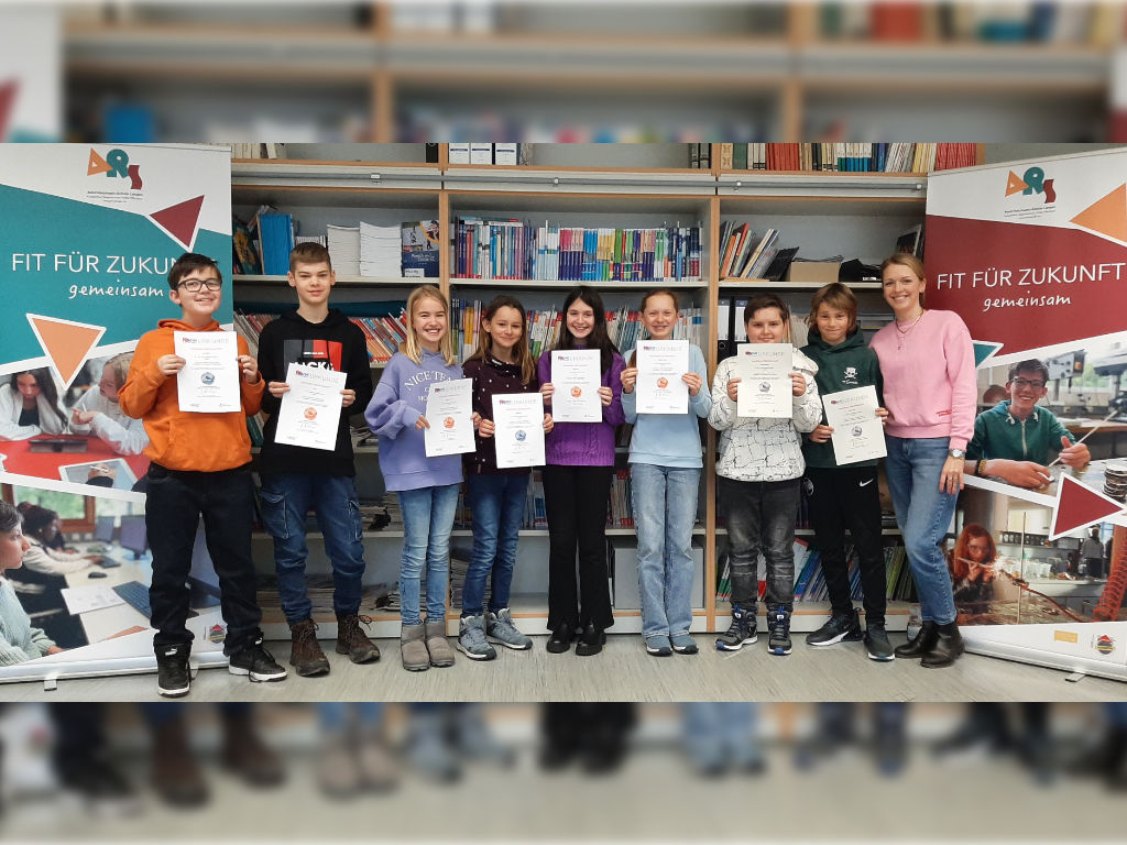 Eine Gruppe Kinder hält Urkunden in die Kamera, dazu eine Lehrkraft, in der Schulbücherei zwischen zwei ARS-Roll-ups mit der Aufschrift „Fit für Zukunft“.
