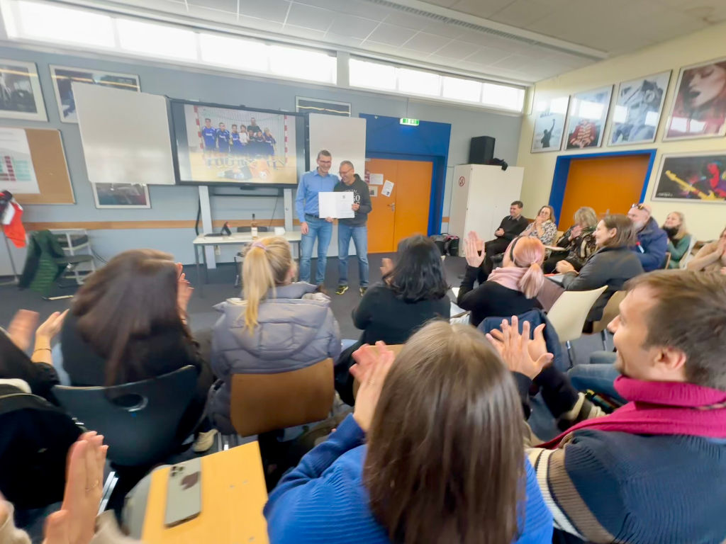 Zwei Männer stehen vorne in einem Raum und halten gemeinsam eine Urkunde; das sitzende Kollegium applaudiert. An den Wänden hängen gerahmte Fotos.