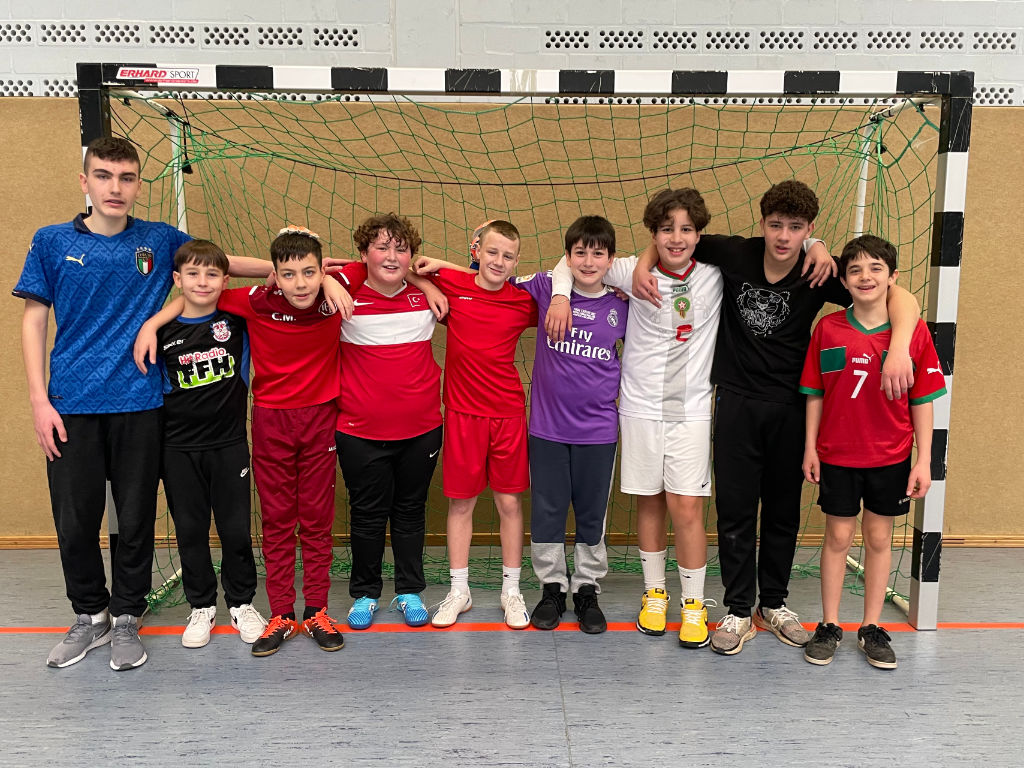 Neun Jungen in verschiedenen Fußballtrikots stehen Arm in Arm vor einem Hallentor in einer Sporthalle.