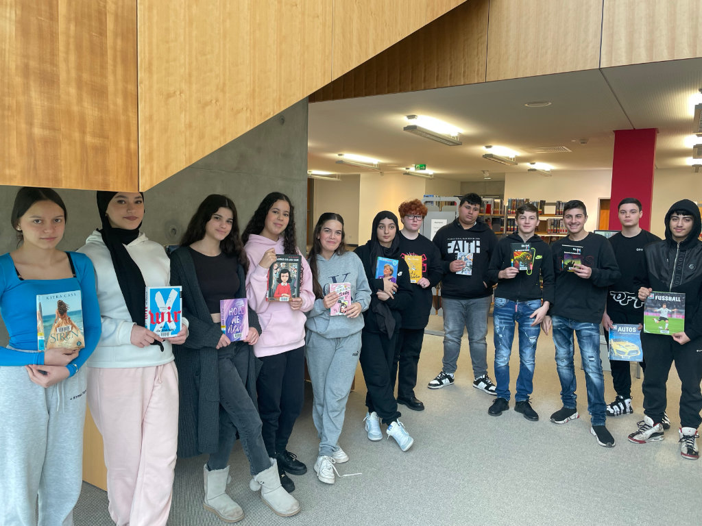 Eine Schulklasse steht in einer Reihe in einer Bücherei; jede Person hält ein Lieblingsbuch in der Hand, im Hintergrund Bücherregale.