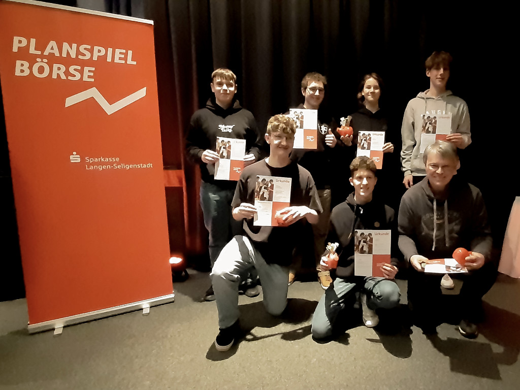 Eine Gruppe Jugendlicher hält Urkunden in die Kamera, dazu eine erwachsene Person, neben einem roten Roll-up mit der Aufschrift „Planspiel Börse – Sparkasse Langen-Seligenstadt“.