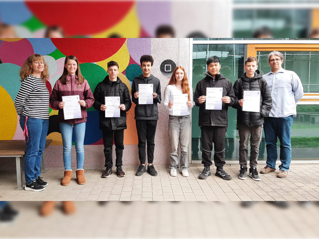 Sechs Jugendliche halten Urkunden in die Kamera, an den Seiten zwei Lehrkräfte, vor einer bunt mit Kreisen bemalten Wand.