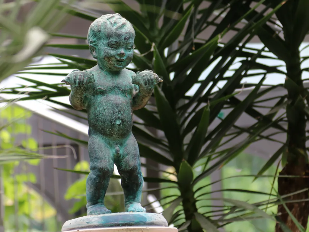 Bronzefigur eines Kindes zwischen Palmwedeln im Gewächshaus.