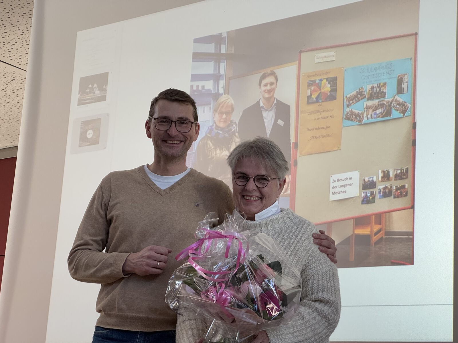 Auf dem Foto stehen zwei Erwachsene im hellen Innenraum der Adolf‑Reichwein‑Schule vor einer großen Projektionsfläche. Links steht der Schulleiter, ein Mann mit kurzen Haaren und Brille, in einem hellen Pullover über einem weißen T‑Shirt. Rechts steht eine Frau mit kurzen, hellgrauen Haaren und runder Brille; sie hält einen Blumenstrauß mit rosa Geschenkband und lächelt zur Kamera. Die Szene zeigt den Abschied der Kollegin in den Ruhestand. Im Hintergrund sind auf der Projektionsfläche unscharf ein Foto von mehreren Personen und bunte Pinnwände zu erkennen.