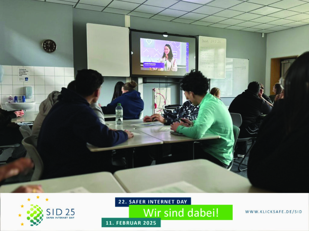 Jugendliche sitzen in einem Klassenraum an Tischen und verfolgen auf einem Bildschirm einen Livestream; unten ein Banner „22. Safer Internet Day – Wir sind dabei!“.