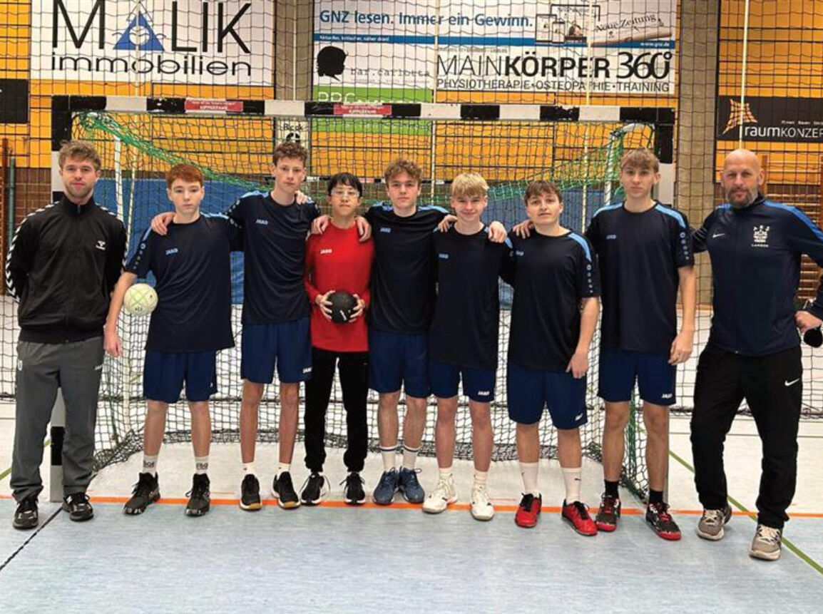 Die Handballmannschaft in dunkelblauen Trikots steht mit zwei Trainern vor einem Hallentor; in der Mitte hält der Torwart in rotem Trikot den Ball.