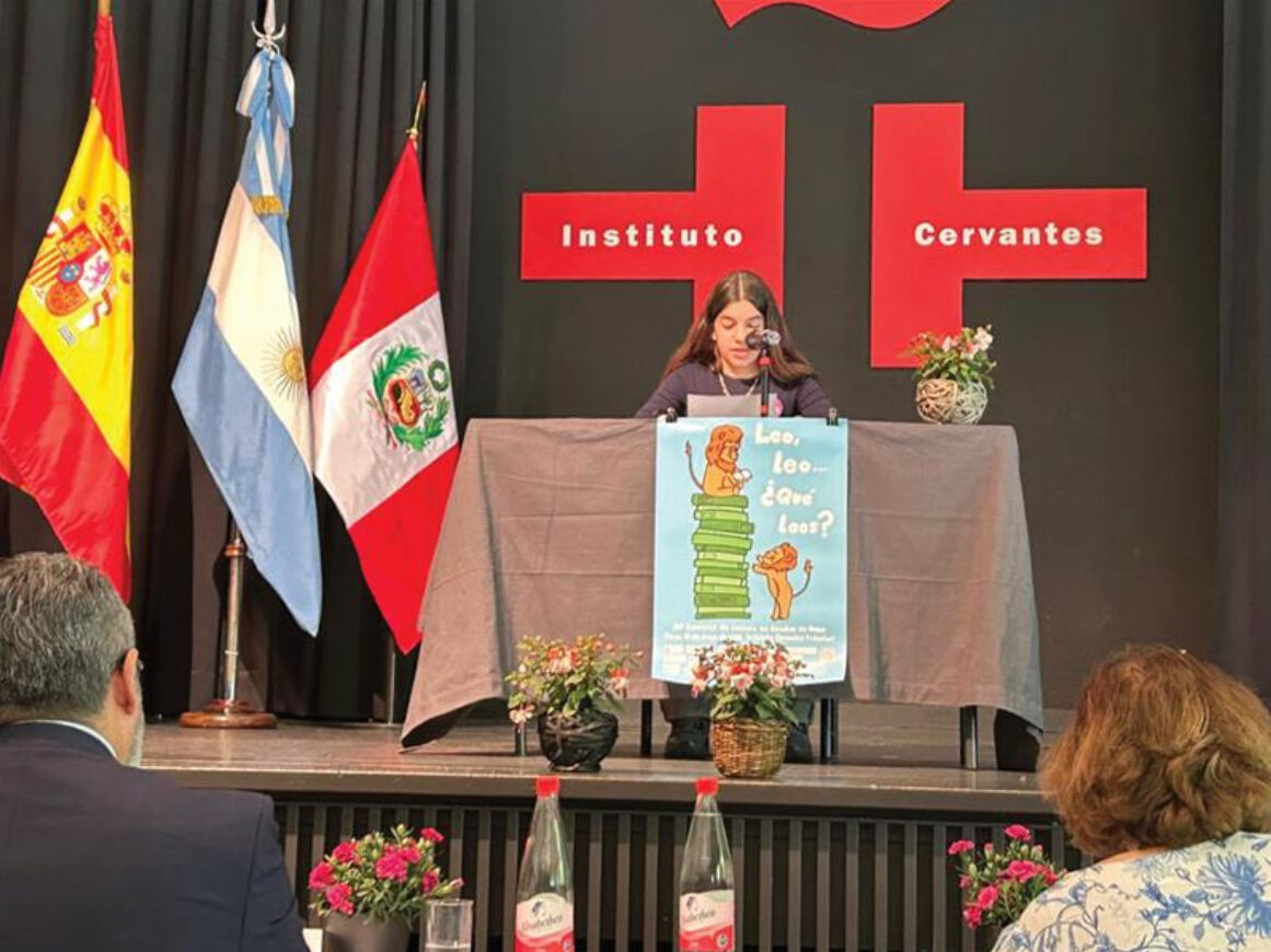 Eine vorlesende Person sitzt an einem Mikrofon auf der Bühne vor der Kulisse des Instituto Cervantes; links stehen die Flaggen Spaniens, Argentiniens und Perus, daneben ein Plakat „Leo, leo… ¿Qué lees?“.