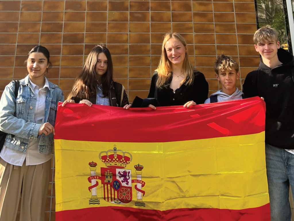 Fünf Jugendliche halten gemeinsam eine große spanische Flagge mit dem Staatswappen vor einer Gebäudefassade.