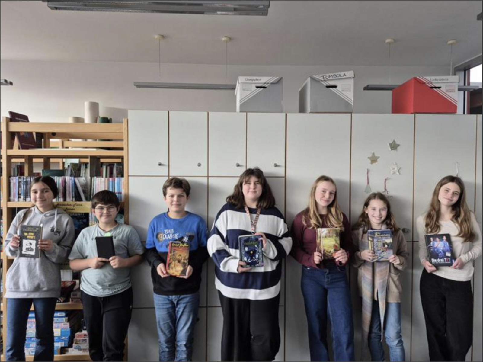 Sieben Kinder stehen nebeneinander in der Schulbücherei und halten jeweils ein Buch in der Hand; im Hintergrund Bücherregale und weiße Schränke.