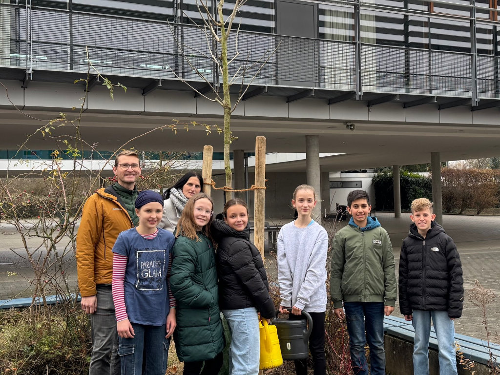 Eine Gruppe Kinder steht mit zwei Erwachsenen neben einem frisch gepflanzten jungen Baum mit Stützpfählen vor dem Schulgebäude; eine gelbe Gießkanne steht bereit.
