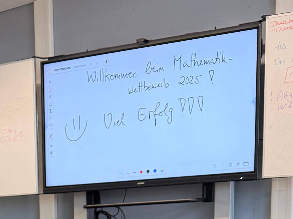 Ein Smartboard mit der handschriftlichen Begrüßung „Willkommen beim Mathematik-Wettbewerb 2025! Viel Erfolg!!!“ und einem gemalten Smiley.