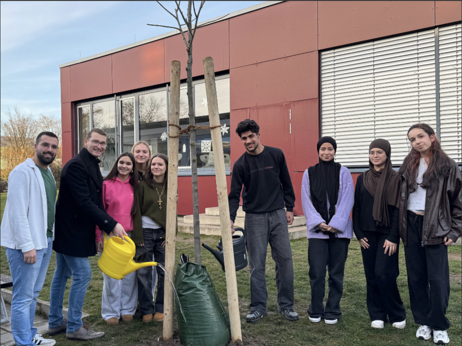 Eine Gruppe Jugendlicher und zwei Lehrkräfte stehen um einen frisch gepflanzten jungen Baum mit Stützpfählen; zwei von ihnen gießen ihn mit Gießkannen.