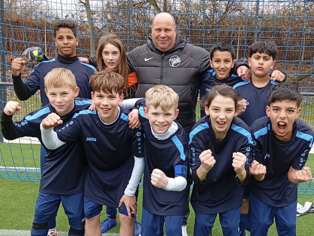 Die Schulfußballmannschaft in dunkelblauen Trikots jubelt gemeinsam mit ihrem Trainer auf dem Sportplatz mit erhobenen Fäusten.