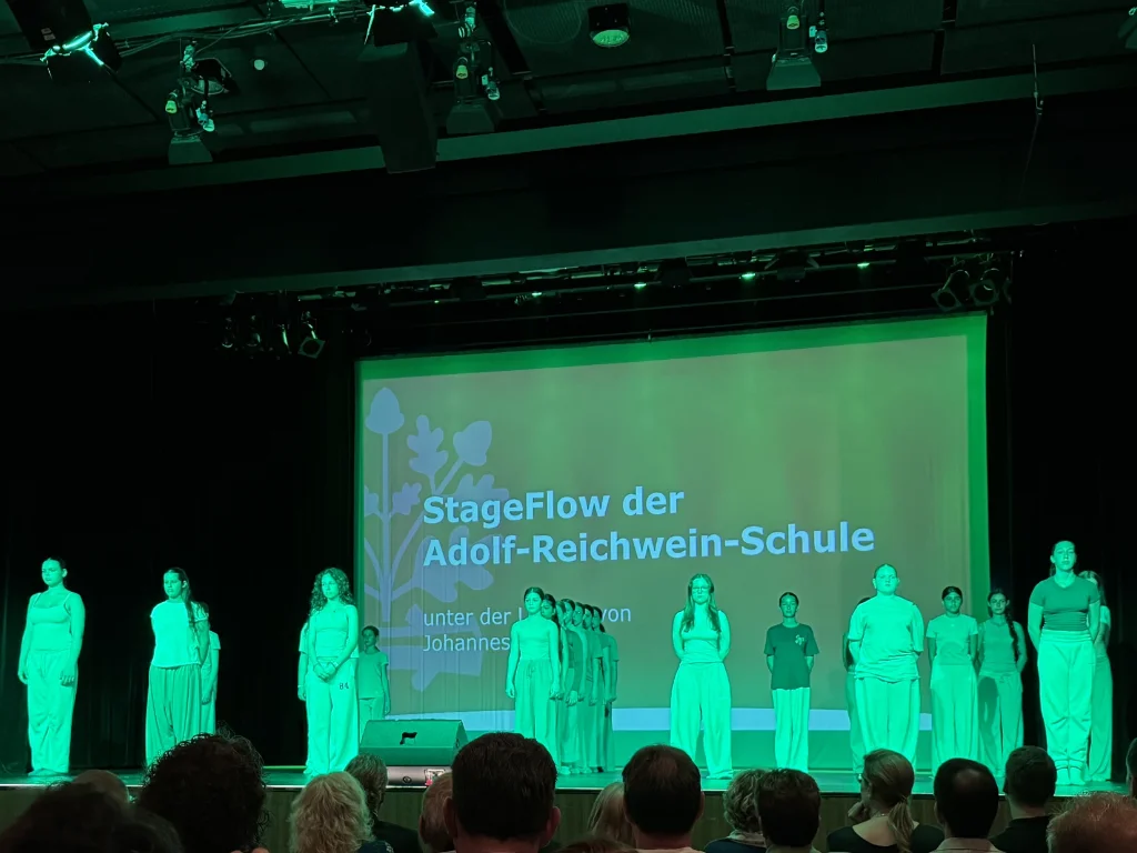 Die Tanzgruppe StageFlow steht in grünem Bühnenlicht auf der Bühne der Stadthalle Langen; im Hintergrund eine Projektion mit dem Titel „StageFlow der Adolf-Reichwein-Schule“.