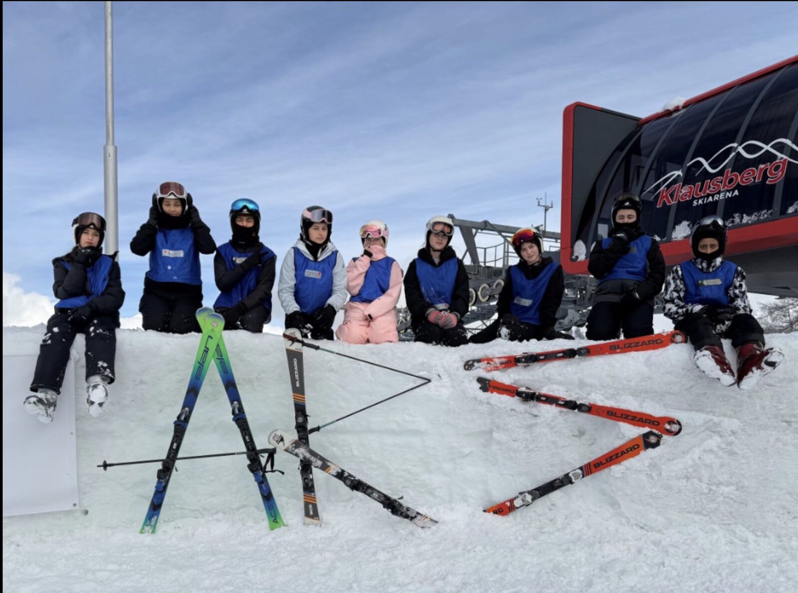 Eine Gruppe Jugendlicher in Skikleidung mit Helm und Skibrille sitzt auf einer Schneemauer im Skigebiet; davor sind Skier und Skistöcke im Schnee zum Schriftzug „ARS“ gelegt.