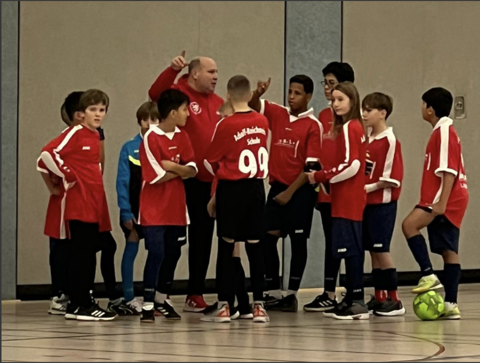 Die Schulfußballmannschaft in roten „Adolf-Reichwein-Schule“-Trikots bespricht sich mit ihrem Trainer in einer Sporthalle.