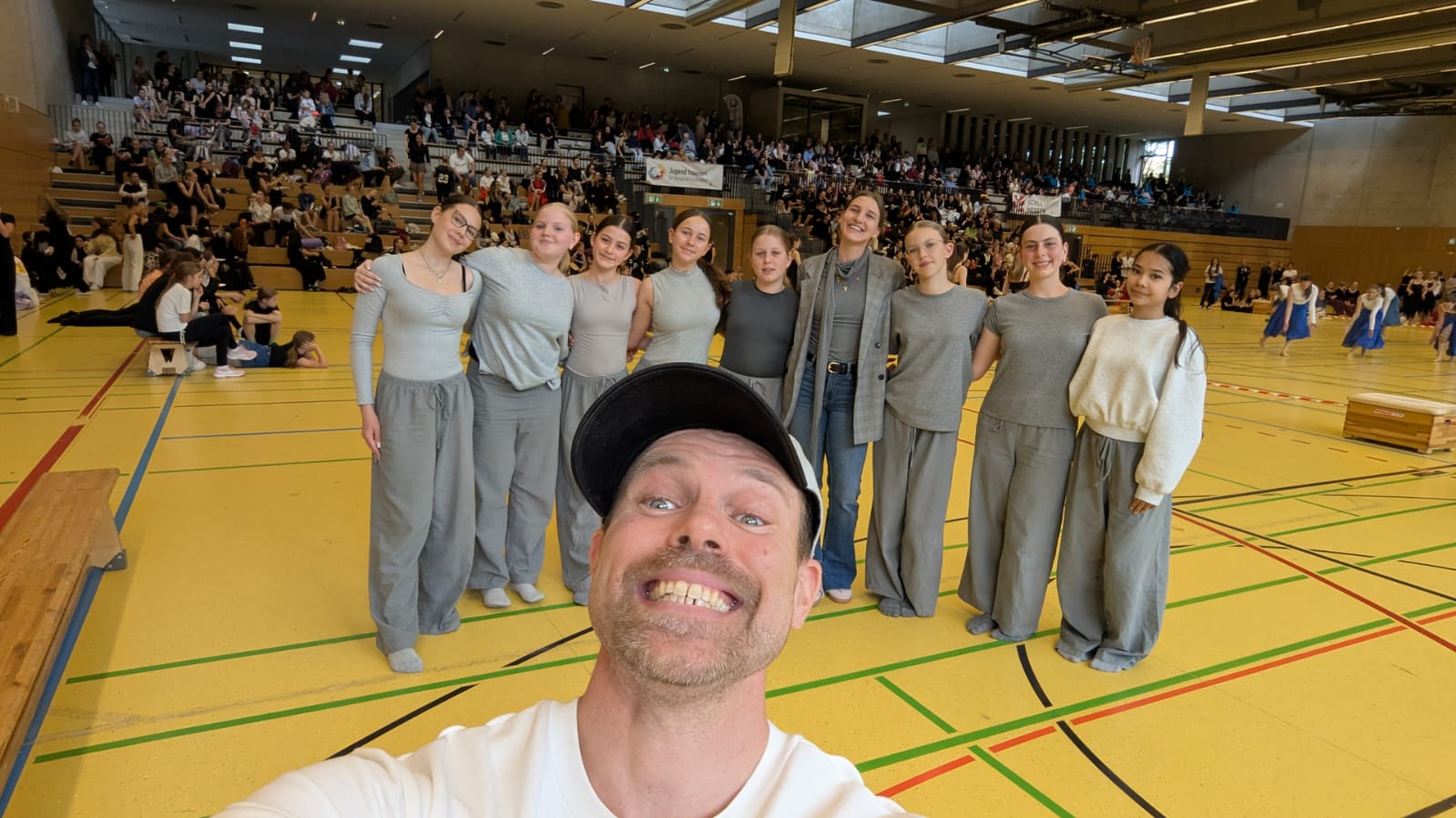 Selfie einer Begleitperson mit der Tanzgruppe StageFlow in einer Sporthalle; auf den Rängen im Hintergrund sitzt zahlreiches Publikum.