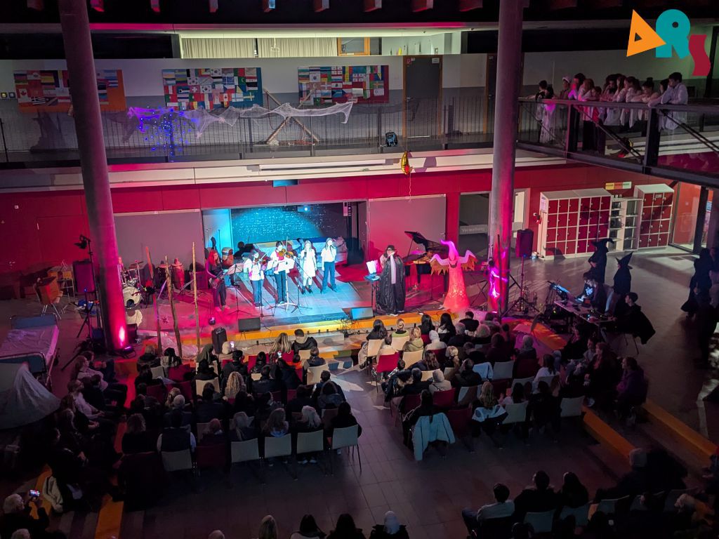 Eine Bühnenaufführung im abendlich rot-violett beleuchteten Schulforum: eine Band und mehrere Singende auf der Bühne, davor ein sitzendes Publikum; ringsum Halloween-Deko wie Spinnweben.