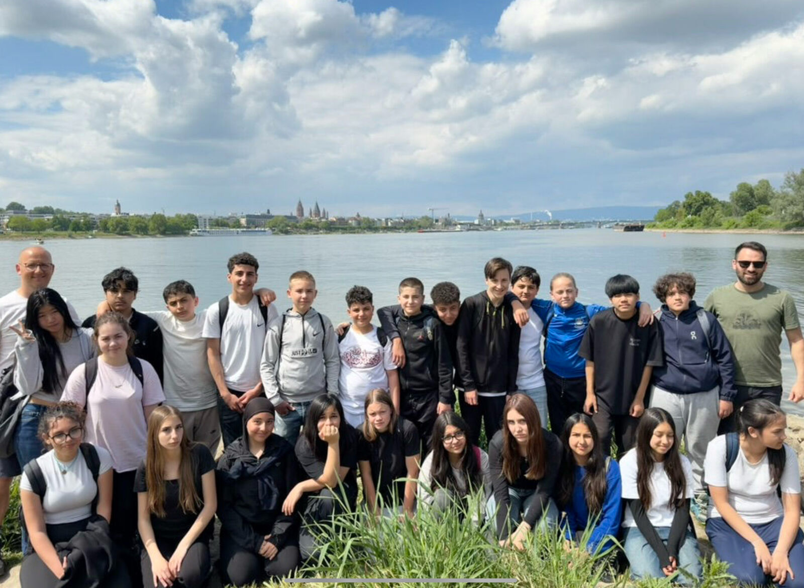 Eine Schulklasse posiert mit zwei Lehrkräften am Ufer des Rheins; auf der gegenüberliegenden Seite ist die Skyline von Mainz mit dem Dom zu sehen.