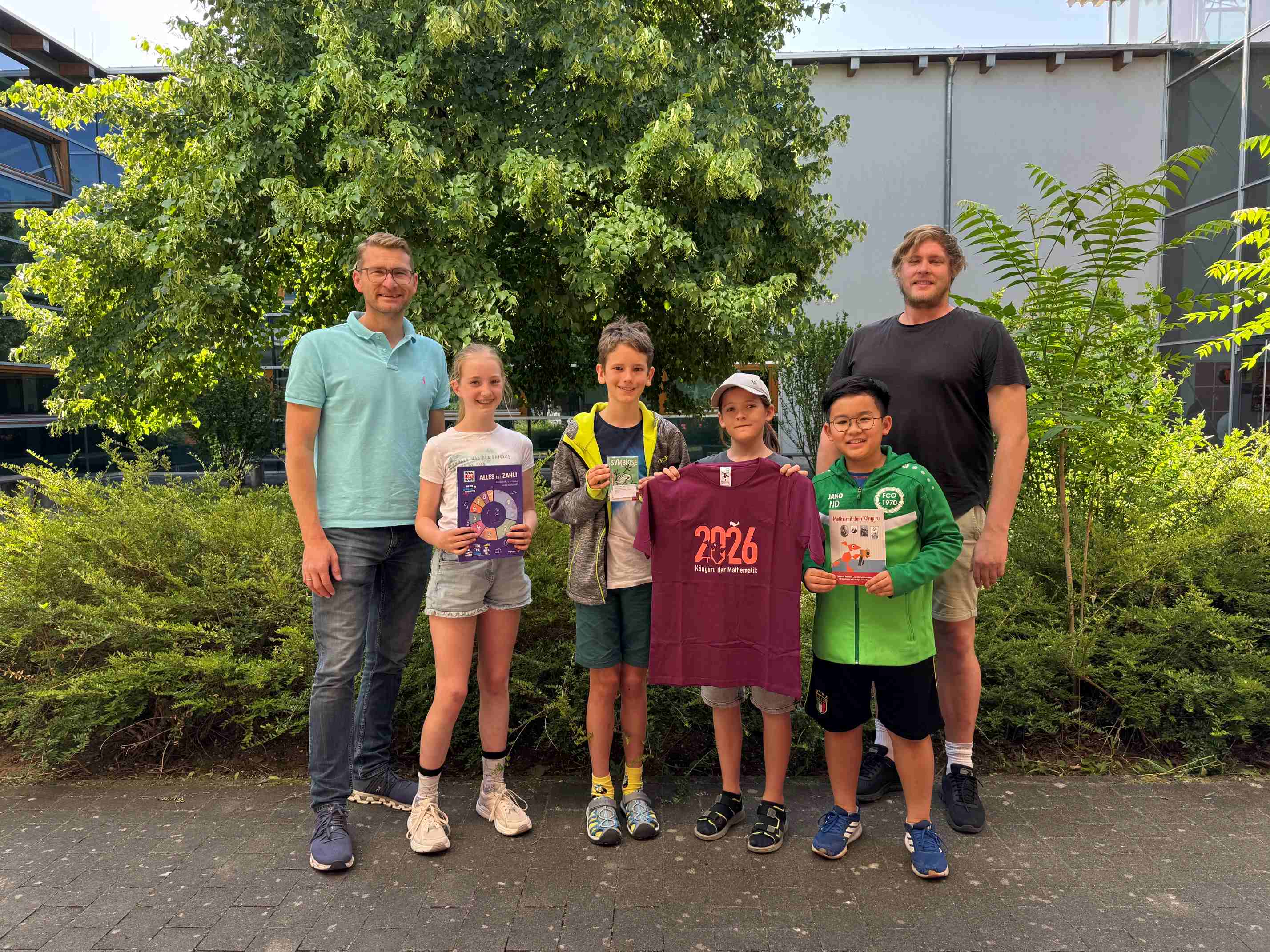 Vier Kinder halten ihre Preise vom Känguru-Wettbewerb in die Höhe – darunter ein T-Shirt mit dem Aufdruck „2026 Känguru der Mathematik“ und Knobelspiele; daneben stehen zwei Lehrkräfte vor Bäumen und dem Schulgebäude.