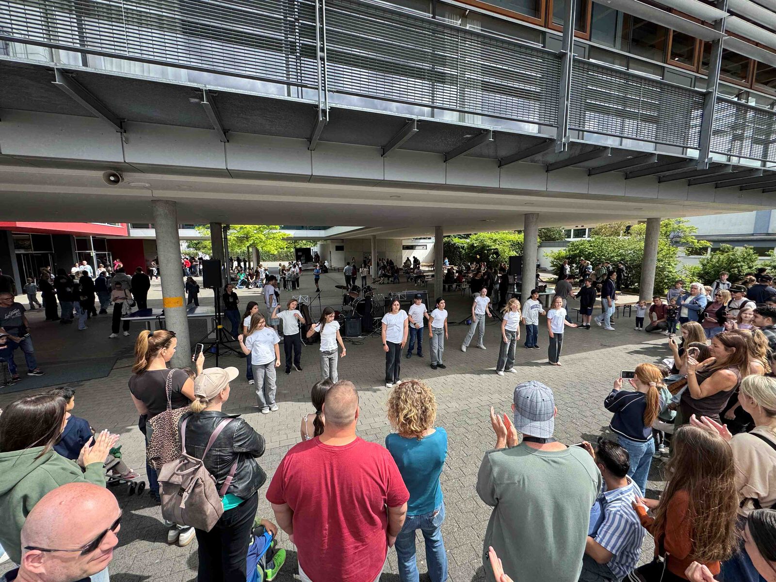 Eine Schülergruppe in weißen T-Shirts führt im Schulhof einen Tanz auf, umringt von zuschauenden und applaudierenden Erwachsenen.