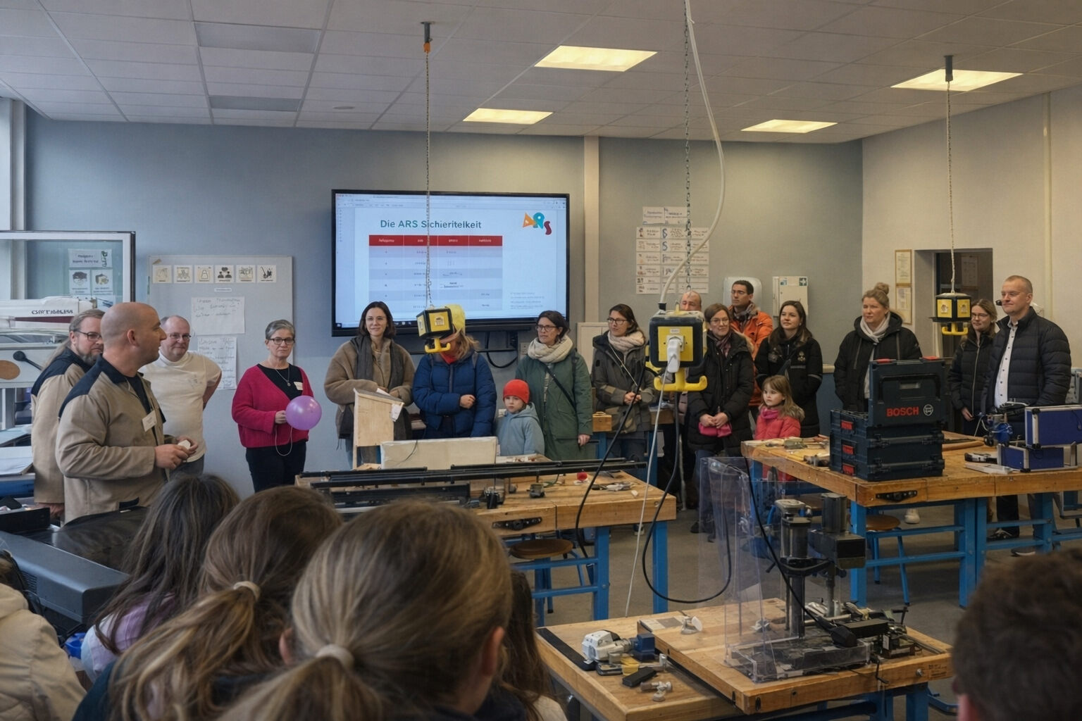 Mehrere Personen stehen im Werkraum der Schule zwischen Werkbänken und Maschinen und verfolgen eine Vorführung; auf dem Smartboard ist eine Übersicht der Schule zu sehen.