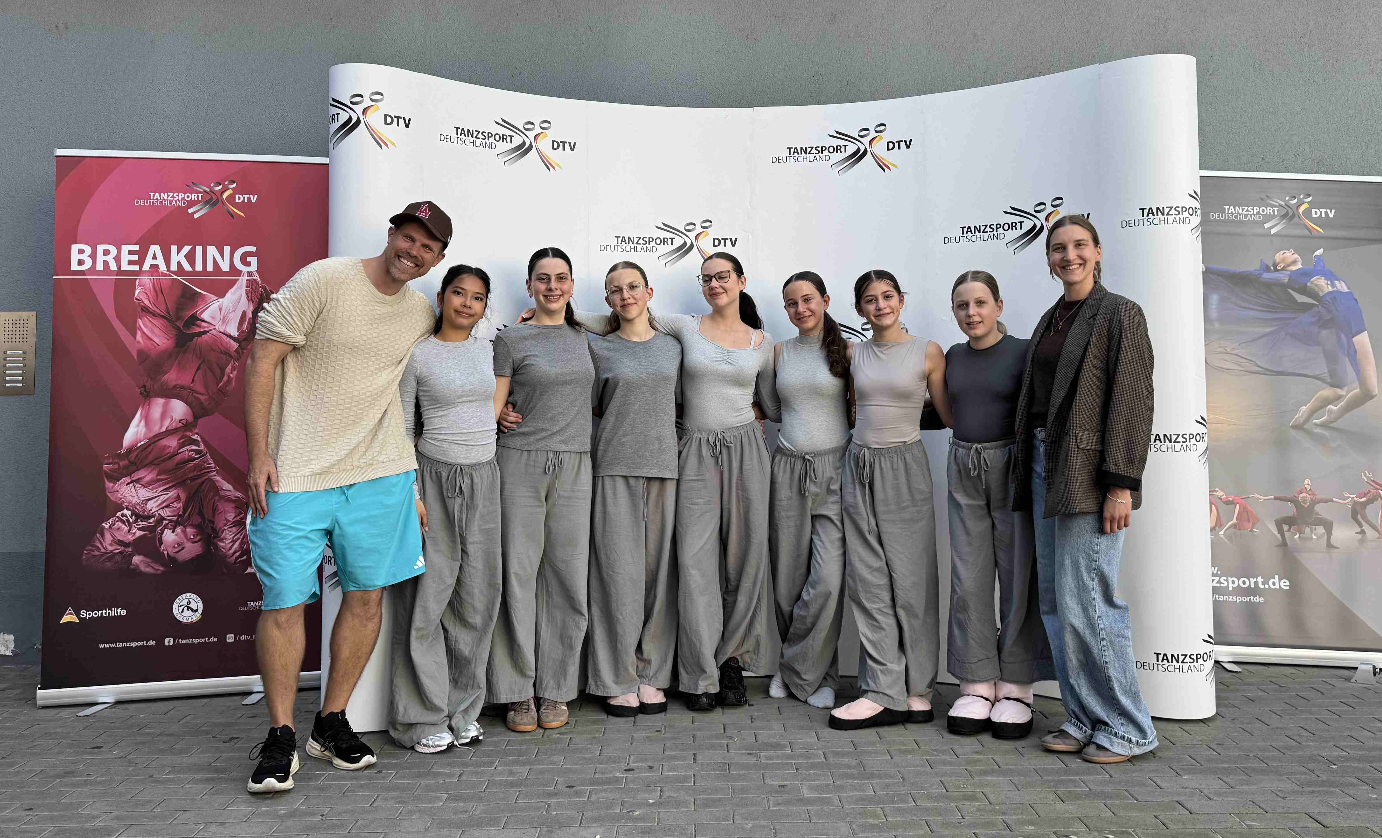 Die Tanzgruppe StageFlow in einheitlichen grauen Outfits sowie zwei Begleitpersonen posieren vor einer Fotowand mit dem Logo „Tanzsport Deutschland DTV“.