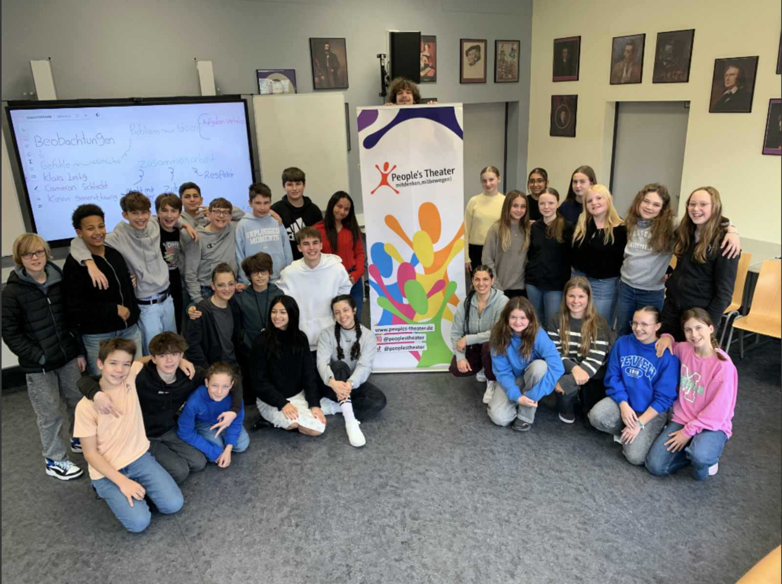 Eine ganze Schulklasse steht und kniet um ein Roll-up mit der Aufschrift „People´s Theater“; im Hintergrund ein Smartboard mit Notizen wie „Respekt“ und „Zusammenarbeit“.