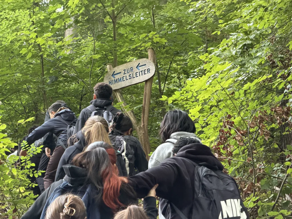 Ein hölzerner Wegweiser mit der Aufschrift „Zur Himmelsleiter“ und zwei blauen Pfeilen steht im grünen Laubwald. Darunter schiebt sich eine Gruppe Jugendlicher mit Rucksäcken den Hang hinauf.