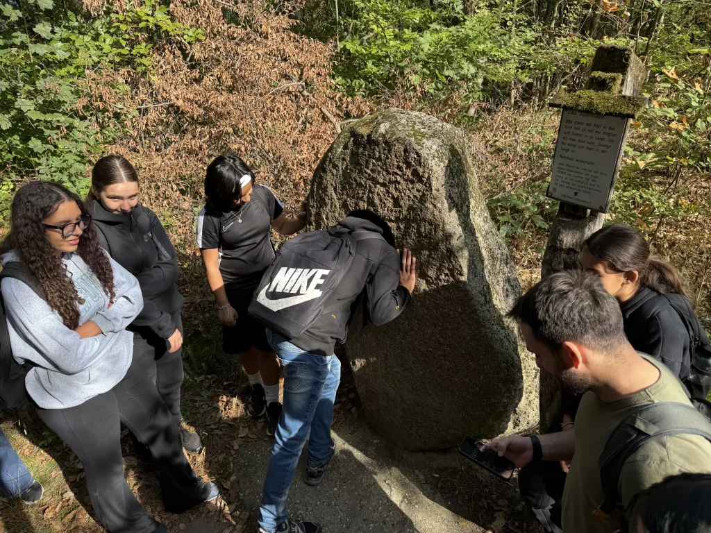 Ein Schüler presst sein Gesicht in die Öffnung eines großen Felsblocks am Wegrand und summt hinein. Mitschülerinnen und Mitschüler stehen im Halbkreis darum herum, einer stoppt mit dem Handy die Zeit. Rechts hängt eine bemooste Informationstafel.