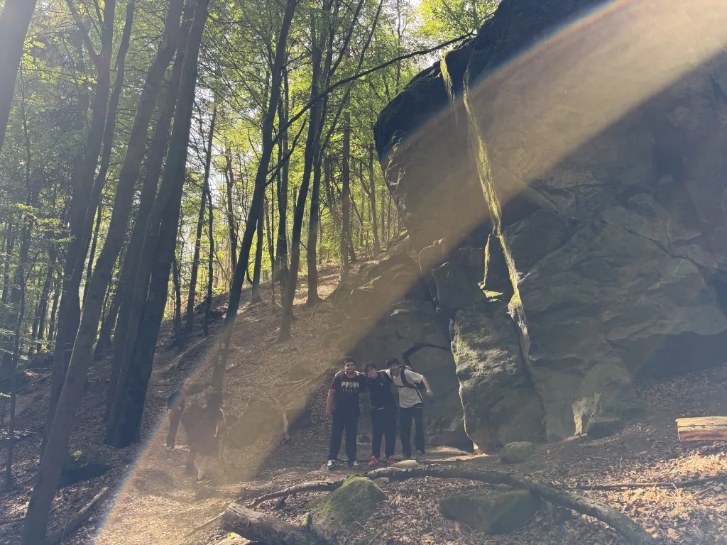 Drei Schüler stehen klein am Fuß eines mehrere Meter hohen, überhängenden Felsblocks. Schräges Sonnenlicht zieht als heller Streifen durch das Bild.