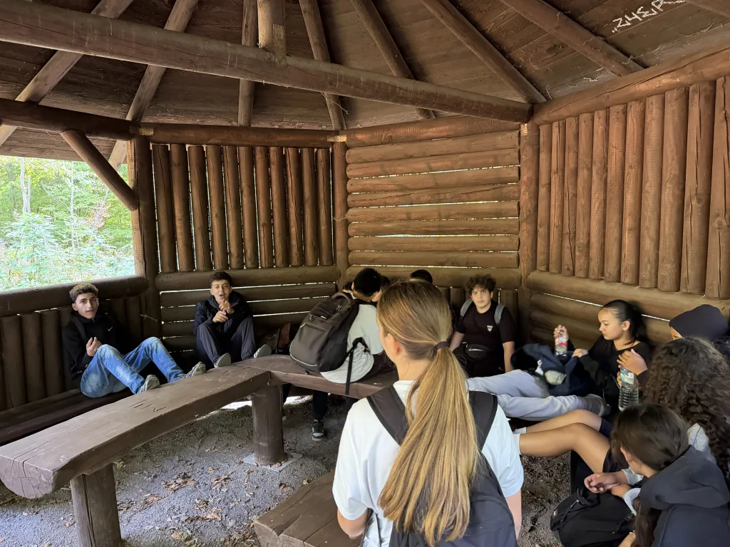 Jugendliche sitzen auf den Holzbänken einer offenen Schutzhütte aus Rundhölzern und unterhalten sich. Durch die offene Seite fällt der Blick in den grünen Wald.