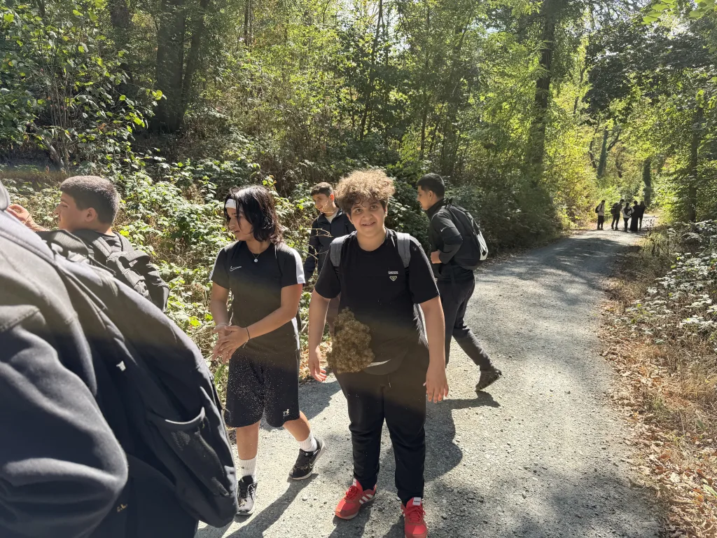 Ein Schüler geht lächelnd über einen breiten Schotterweg, an seinem schwarzen T-Shirt klebt ein Ball aus Kletten. Neben und hinter ihm gehen weitere Jugendliche.