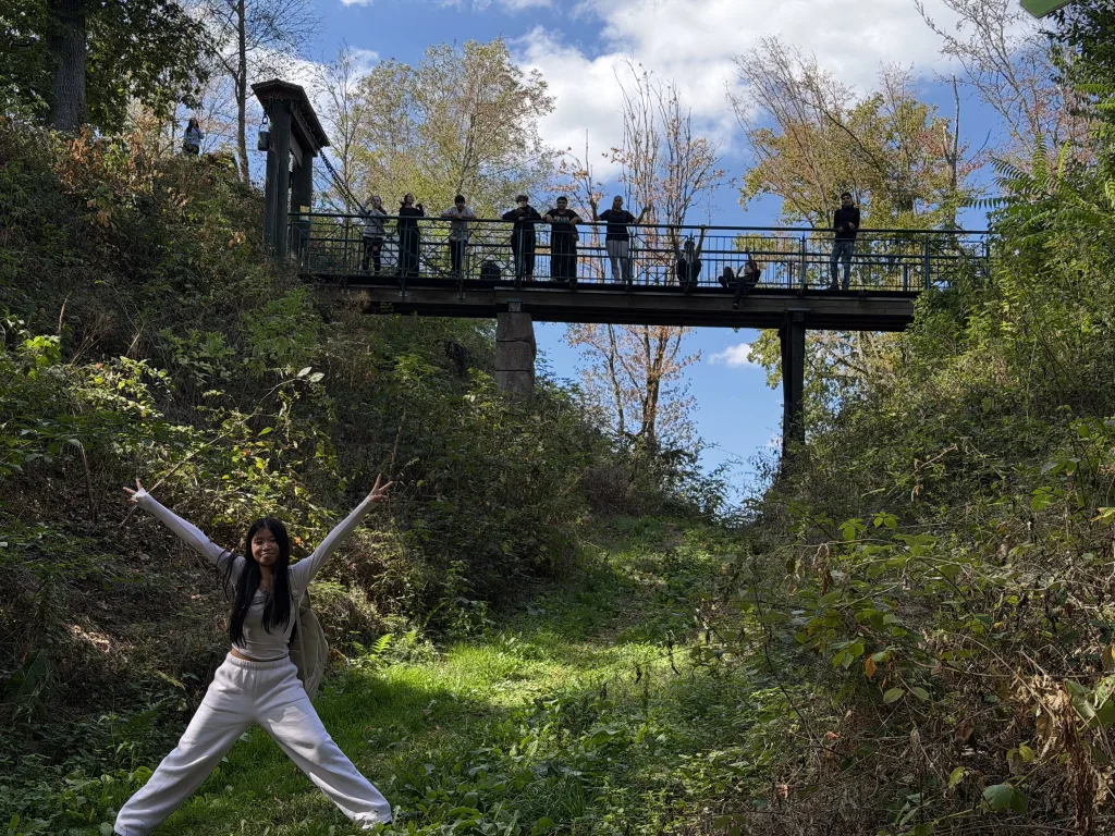 Eine eiserne Fußgängerbrücke überspannt eine grüne Senke, oben stehen Jugendliche am Geländer. Unten auf der Wiese posiert eine Schülerin mit ausgebreiteten Armen.