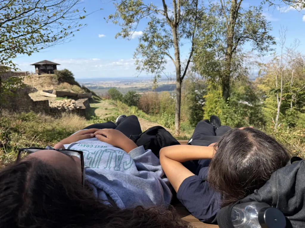 Aus liegender Perspektive fotografiert: Drei Jugendliche liegen auf einer Bank. Dahinter stehen eine Schutzhütte und eine alte Mauer, im Hintergrund die weite, flache Landschaft unter blauem Himmel.