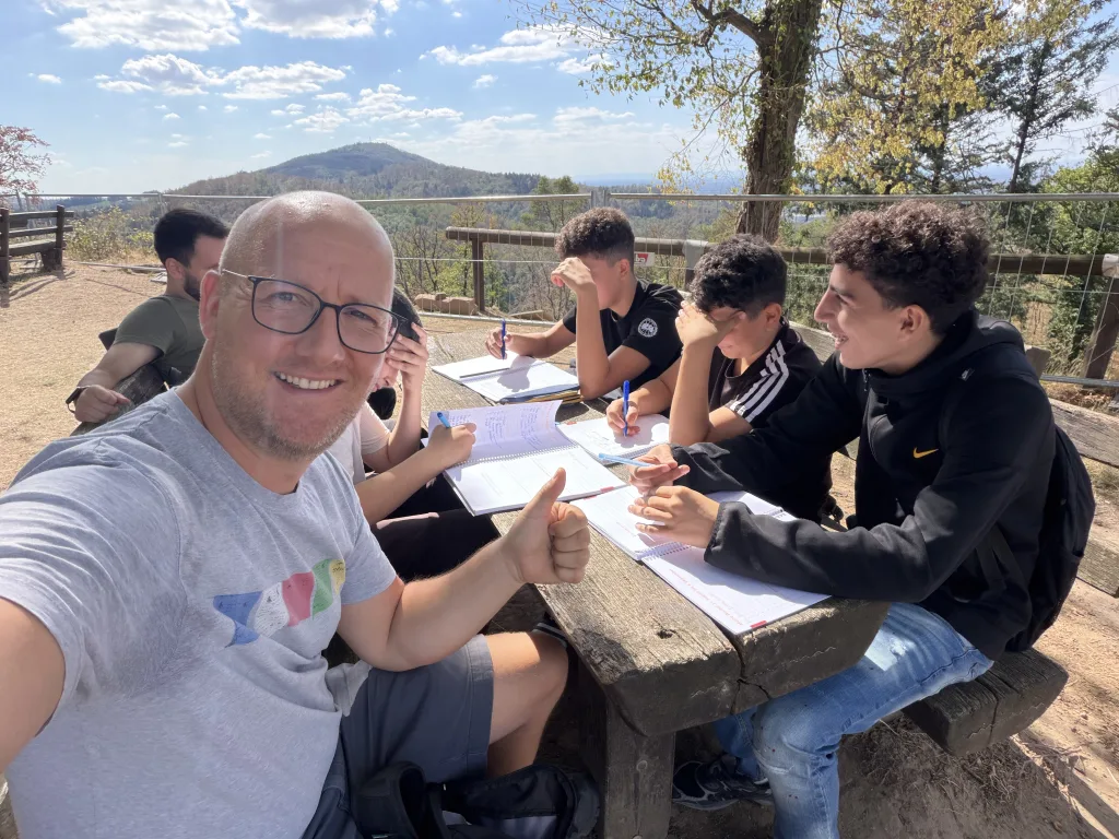 Selfie eines Lehrers mit Brille, der lächelt und den Daumen hebt. Hinter ihm sitzen vier Schüler an einem Holztisch und schreiben in ihre Logbücher. Im Hintergrund ein bewaldeter Berg und blauer Himmel.
