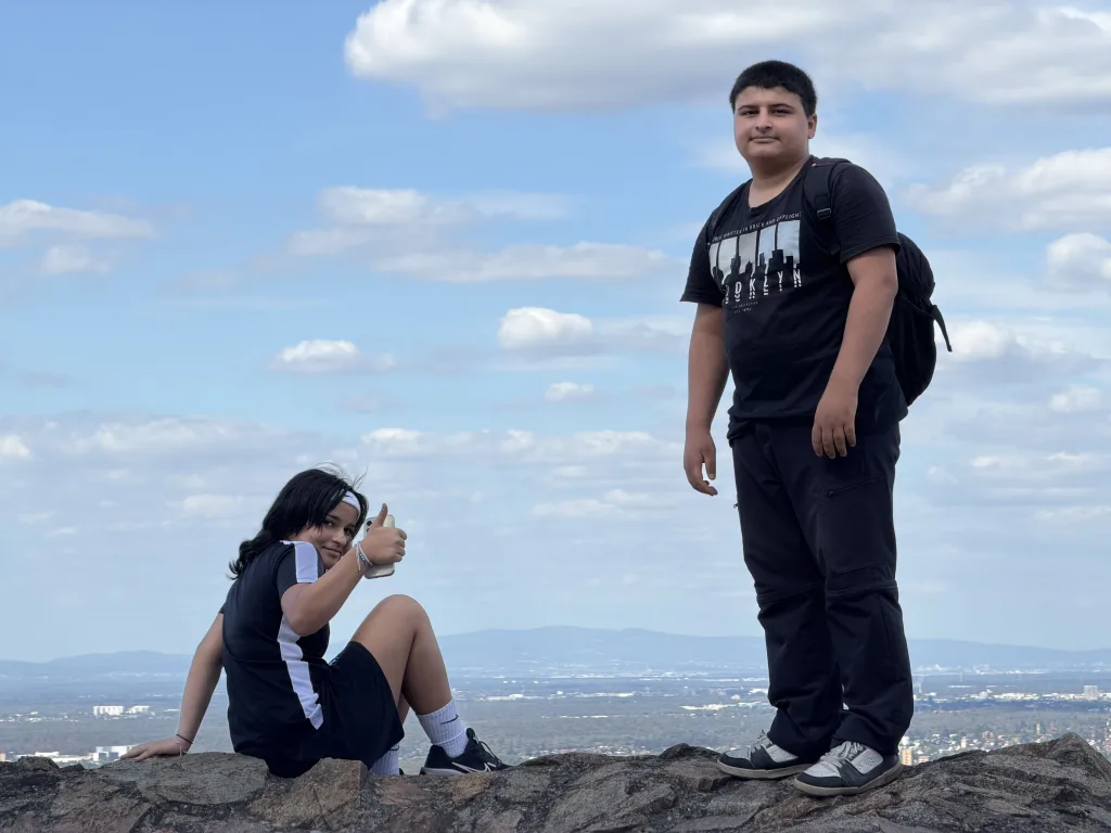 Eine Schülerin sitzt auf einer Felsmauer und zeigt Daumen hoch, daneben steht ein Schüler mit Rucksack. Hinter beiden nur Himmel mit Schäfchenwolken und die Ebene bis zum Horizont.