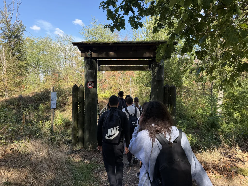 Eine Gruppe geht durch ein hölzernes Tor mit Giebeldach, das den Ausgang der Burgruine bildet. Dahinter führt der Weg in den Wald hinab.