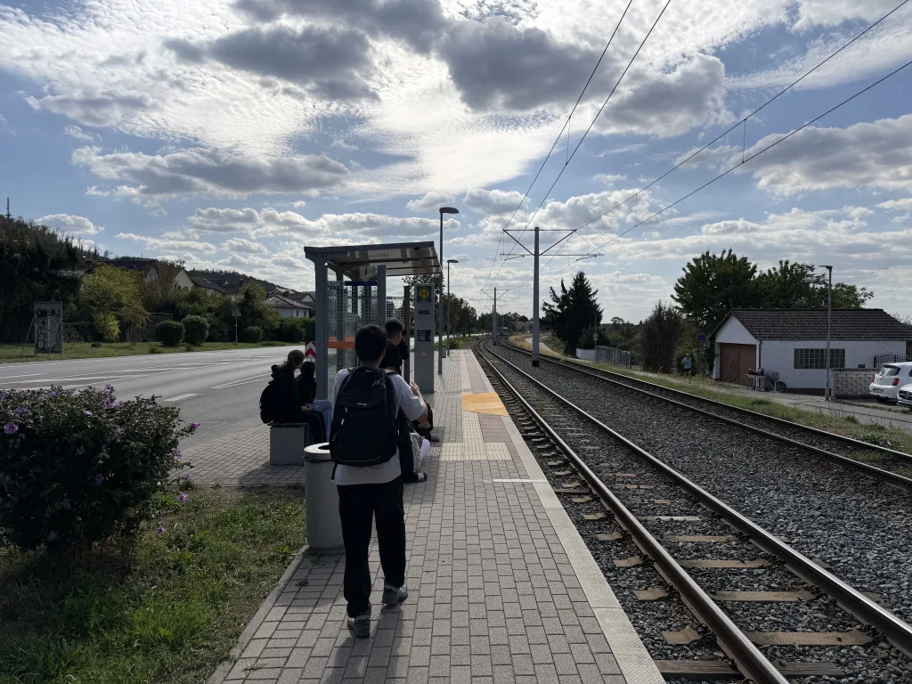 Ein Bahnsteig mit Wartehäuschen neben zwei Gleisen, ein Schüler mit Rucksack geht darauf zu. Weiter vorn sitzen einige auf der Bank, darüber ein weiter Wolkenhimmel.