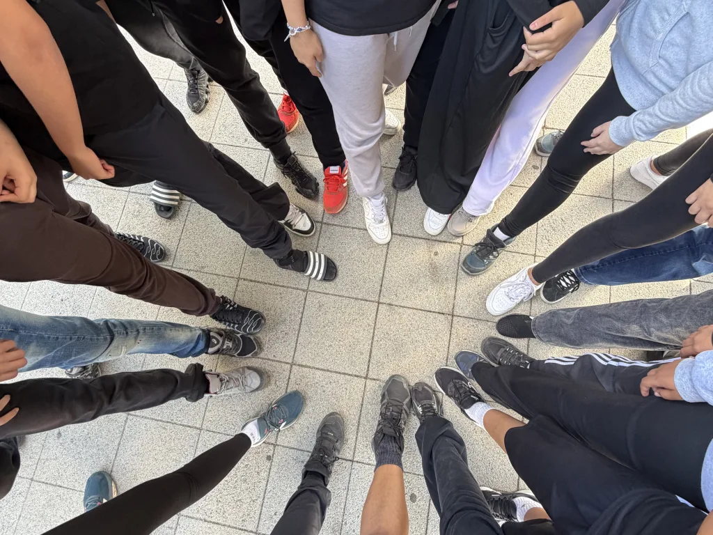 Blick von oben auf einen Kreis aus Füßen auf hellen Bodenplatten: überwiegend Sneaker und Turnschuhe, dazu ein Paar feste Wanderstiefel und ein Paar schwarze Badelatschen mit drei weißen Streifen.