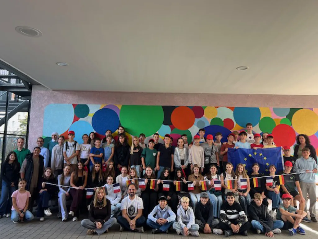Eine große Gruppe von Schülerinnen und Schülern sowie Lehrkräften steht gemeinsam in einem Schulgebäude für ein Gruppenfoto. Im Hintergrund ist eine farbenfrohe Wandgestaltung mit großen bunten Kreisen zu sehen. In der Mitte der Gruppe wird eine Europaflagge gehalten. Vor der Gruppe hängen kleine Flaggen verschiedener europäischer Länder an einer Schnur. Die Teilnehmerinnen und Teilnehmer stehen und sitzen in mehreren Reihen und blicken in die Kamera.